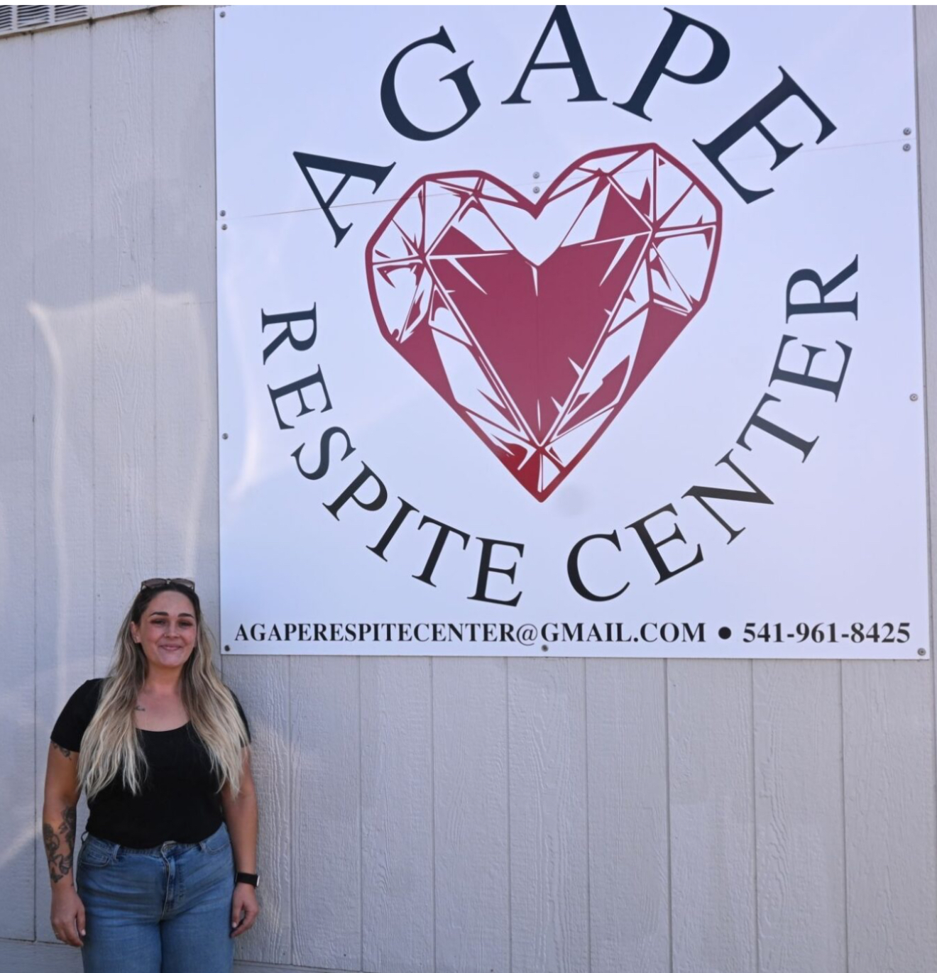 The Agape Respite Center storefront in Newport, Oregon, with the executive director standing in front of a large white-and-navy sign featuring a ruby heart logo and the words 'Agape Respite Center.'
