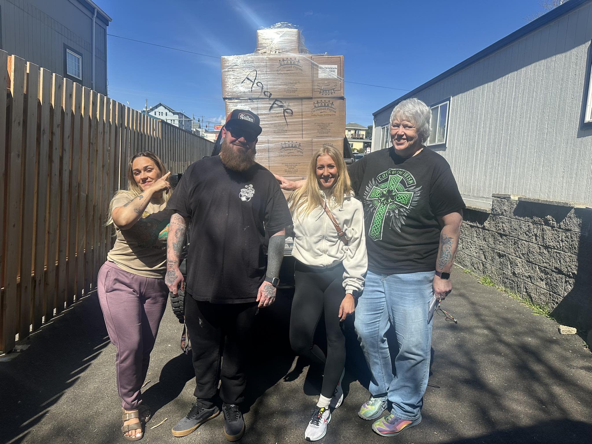 Four Agape Respite Center volunteers smiling in front of a tall, shrink-wrapped pallet of Agape donation boxes outside the center in Newport, Oregon